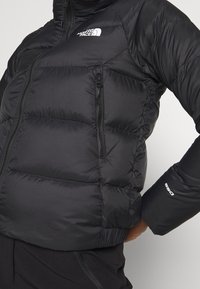 Doudoune noire avec un design matelassé, poche poitrine zippée et poignets élastiques. Caractéristiques d'un col montant et du logo The North Face.