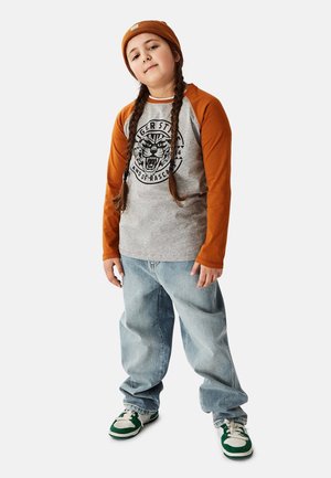 Langærmet grå T-shirt med orange raglansærmer, med et sort tigerprint. Kombineret med løstsiddende lyseblå jeans og grønne sneakers.