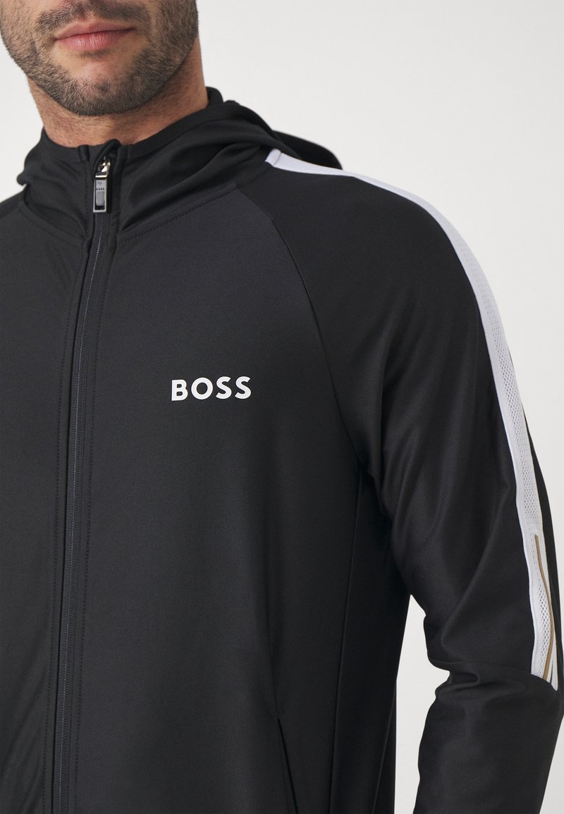 Schwarze Zip-Jacke mit Kapuze, ausgestattet mit weißen Seitenelementen und dem BOSS-Logo auf der Vorderseite. Hergestellt aus glattem, dehnbarem Material mit einem figurbetonten Design.