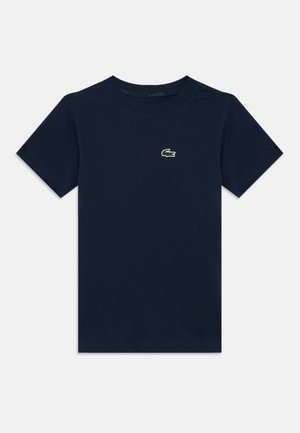 T-shirt de algodão azul marinho com mangas curtas, apresentando um pequeno logo da Lacoste em verde e vermelho no peito. Gola simples em formato de barco.