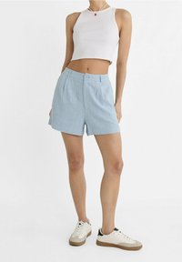 Pantaloni corti azzurri in una vestibilità rilassata con frontale piatto, pince e chiusura con un bottone. Indossati con un top corto bianco e sneakers.