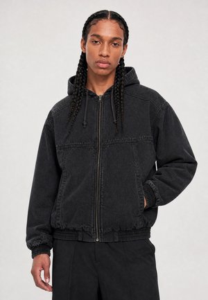 Jeune personne aux longs cheveux tressés portant un sweat à capuche zippé noir et un pantalon noir, debout avec une main dans la poche sur un fond uni.