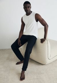 White sleeveless top paired with black jogger pants, seated on a textured, light-colored chair in a minimalistic indoor setting.