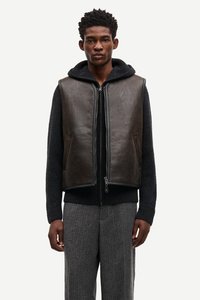 Temno rjava usnjena jopica preko črne pletenine s kapuco, z zadrgo in dvema sprednjima žepoma, v kombinaciji s sivimi teksturiranimi hlačami.