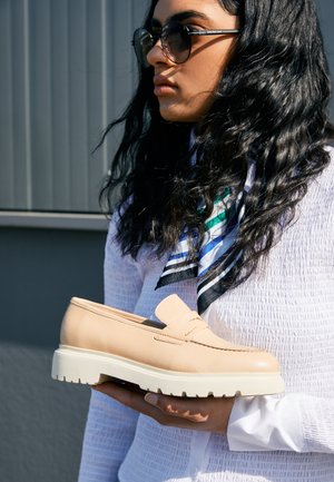 Woman with long dark hair and sunglasses holds beige chunky-soled loafer against gray background, wearing white textured shirt and striped scarf.