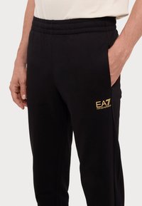 Fekete melegítőnadrág, laza szabással, rugalmas derékkal, oldalsó zsebekkel és arany EA7 logóval a bal combon.