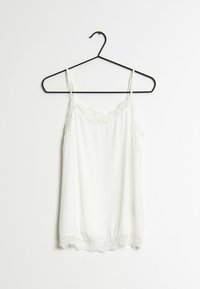 Vit linne hängande på en svart krok, med spetsdetaljer vid halsringningen och fållen, samt tunna justerbara band och en slät yta.