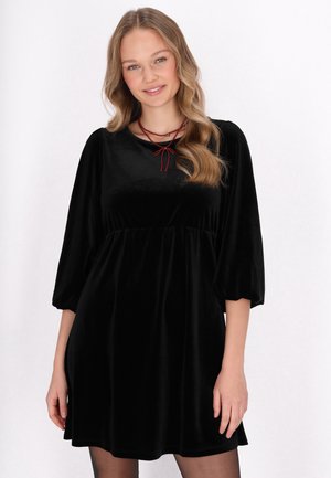 Rochie din catifea neagră cu mâneci puff până la trei sferturi și talie strânsă. Prezintă un colier din sfoară roșie legat la gât.