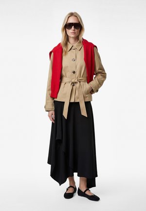 Frau trägt übergroße Sonnenbrille, beige Jacke mit Gürtel, roten Pullover über die Schultern gelegt, schwarzen asymmetrischen Rock und schwarze Ballerinas.