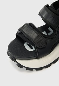 Sandalha preta com tiras ajustáveis em velcro, palmilha texturizada e uma sola branca grossa. Apresenta detalhes com o logótipo e um padrão de piso robusto.