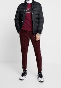 Svart puffjacka över en vinröd sweatshirt, ihop med vinröda tapered byxor och svart-vita sneakers. Har synlig sömnad och logotypdetaljer.