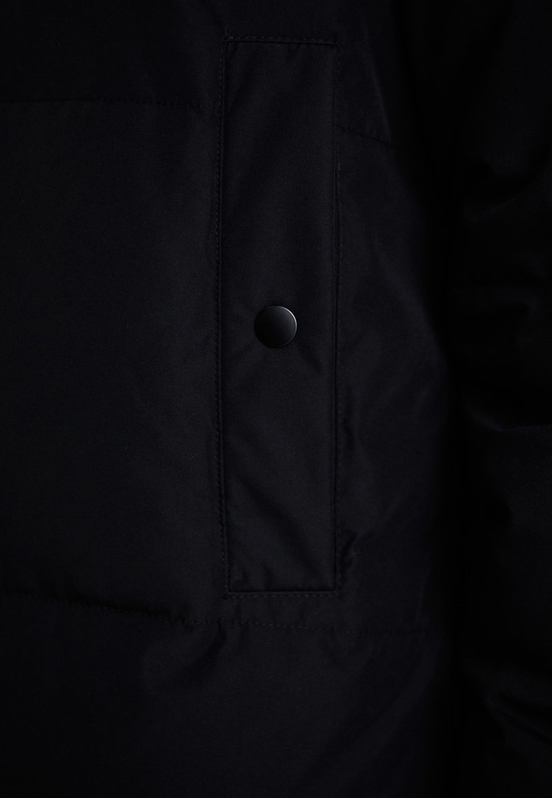 Schwarze Daunenjacke mit mattem Finish, ausgestattet mit einer aufgesetzten Tasche und einem schwarzen Druckknopf. Strukturiertes Gewebe mit dezenten Steppdetails.