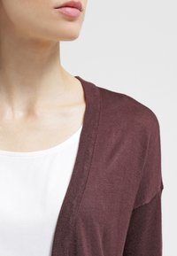 Žena v bílém tričku a hnědém pleteném kardiganu, viditelná od ramen po rty na jednobarevném světlem pozadí.