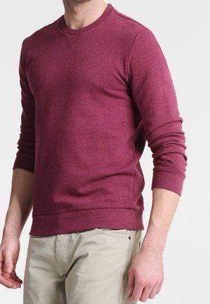 Mann in einem enganliegenden burgunderfarbenen Langarmpullover und beigen Hosen, mit entspannten Händen an den Seiten vor einem weißen Hintergrund.