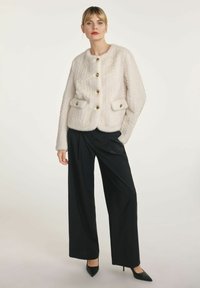 Biała futrzana kurtka z teksturowanym wzorem, okrągłym kołnierzem i złotymi guzikami, zestawiona z czarnymi spodniami w szerokim kroju i spiczastymi szpilkami.