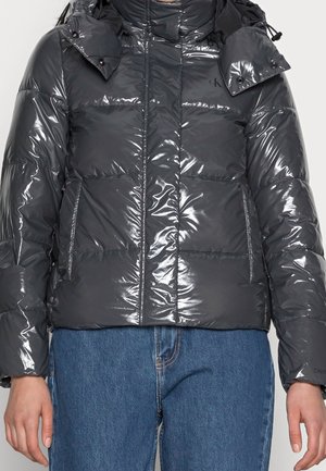 Skinnende sort dunjakke med hætte og lynlåslommer båret over blå jeans, hænder afslappet ned langs siderne.