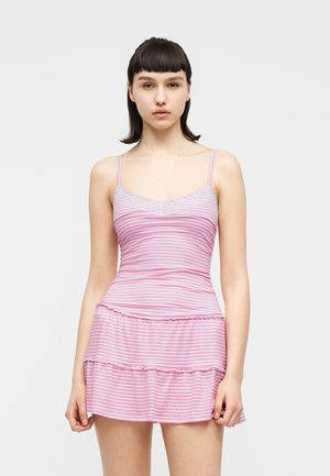 Junge Frau mit glattem dunklem Haar, die ein ärmelloses, pink-weiß gestreiftes Kleid mit Spitzenbesatz trägt und vor einem schlichten Hintergrund steht.