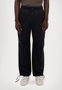 Man wearing black loose-fit cargo pants with drawstrings at the ankles and white sneakers, standing against a plain background.