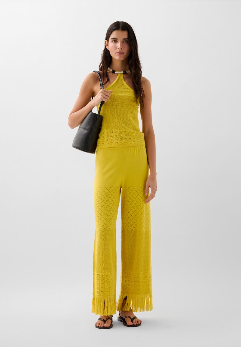 Débardeur en maille jaune avec cou à halter, associé à un pantalon en maille à jambes larges avec un motif texturé et des ourlets à franges. Sac pochette noir inclus.