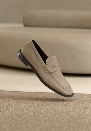 Mocassin en daim beige avec une semelle foncée, présenté sur un fond de tapis et canapé assortis.