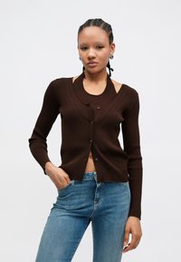 Καφέ πλεκτή ζακέτα με ριγέ υφή, στρογγυλή λαιμόκοψη, κούμπωμα με κουμπιά και σχέδιο με ανοίγματα στους ώμους, σε συνδυασμό με ανοιχτόχρωμο μπλε τζιν.