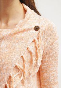 Pull en maille orange et crème avec un col châle, orné d'un détail frangé et d'un bouton marron sur l'épaule.