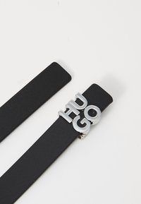 Cintura in pelle nera con texture liscia, caratterizzata da lettere metalliche in argento "HUGO" come accentuata della fibbia. Design semplice e pulito.