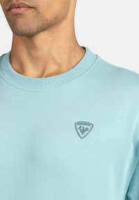 Sudadera azul claro con cuello redondo de canalé y un parche de logo gris en el lado izquierdo del pecho. La tela parece suave y ligera.