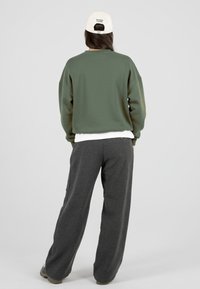 Asmuo, vilkintis baltą kepurę, alyvuogių žalios spalvos megztinį, baltą marškinėlį ir laisvas tamsiai pilkas kelnes, stovi nugara pasukęs į žiūrovą.