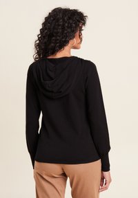 Schwarzer, langärmeliger Hoodie mit geripptem Saum und Bündchen. Hat eine weiche Textur und eine Stoffkapuze, getragen mit beigen Hosen.