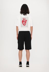 T-shirt bianca oversize con design di un cuore anatomico rosso su sfondo testuale. Abbinata a pantaloni neri e sneaker bianche con accenti neri.