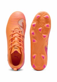 Φωτεινά πορτοκαλί ποδοσφαιρικά παπούτσια με ροζ λεπτομέρειες, που διαθέτουν υφή στην επάνω πλευρά, κλείσιμο με κορδόνια και κωνικά καρφιά στη σόλα για καλύτερη πρόσφυση.