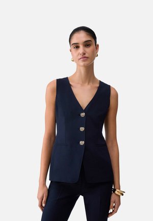 Donna che indossa un gilet senza maniche blu navy con tre bottoni decorativi, abbinato a pantaloni blu navy coordinati e gioielli dorati a polso e orecchie.