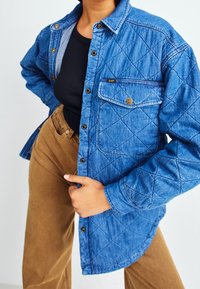 Quiltad blå denimjacka med knappstängning framtill, en bröstficka och gyllene detaljer. Kombinationen med bruna byxor visar en avslappnad stil.
