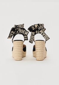 Mustad espadrillid, millel on tekstuuriga jute kontsad, laia rihmaga ja suured mustad ning beežid mustriga kaared pahkluu ümber.