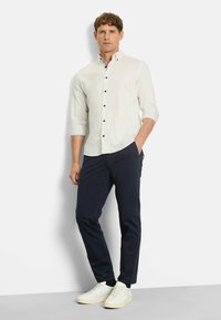 Helles beige Hemd mit Knöpfen, navyfarbene Hose und cremefarbene Sneaker. Das Hemd hat einen umgeschlagenen Kragen und dunkle Knöpfe. Maßgeschneiderter Schnitt.