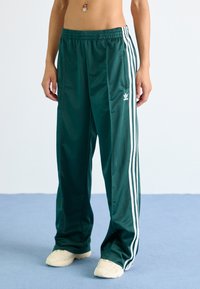 Green, lightweight track pants with white side stripes, elastic waistband, and side pockets. Styled with beige sneakers.