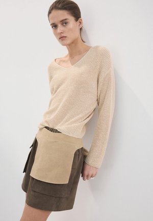 Mladata žena naslonjena na zid, nosi bež pleteni džemper i smeđu suknju od brušene kože s velikim smeđim džepnim pregačem.