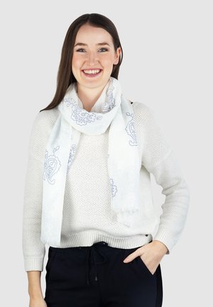 Witte sjaal met blauw bloemenpatroon, lichtgewicht stof, franje randen, gedragen over een wit gebreid sweater. Subtiele ontwerpelementen overal.