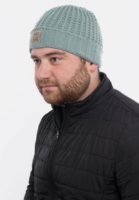 Jasnoniebieska dziana czapka z fakturowanym wzorem i ściągaczem, z brązową skórzaną naszywką z logo. Noszona przez osobę w czarnej kurtce.