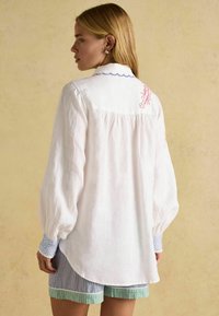 Weißes Leinenhemd mit Puffärmeln, blauem gewelltem Kragen und rotem gesticktem Design auf der Rückseite. Getragen mit gestreiften Shorts.