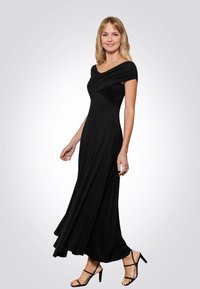 Abito nero lungo fino al pavimento con corpetto aderente, scollatura off-the-shoulder e dettagli arricciati; presenta una gonna fluente e sandali con tacco a blocco.