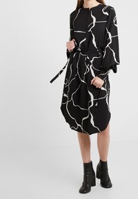 Schwarzes Kleid mit weißen abstrakten Linien, langen Ärmeln, runder Ausschnitt und Taillengürtel; strukturierter Stoff mit lockerer Silhouette.