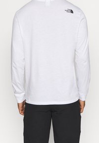 Vit långärmad t-shirt i bomull, med en liten svart logotyp på övre delen av ryggen, rund halsringning och åtsittande ärmar.