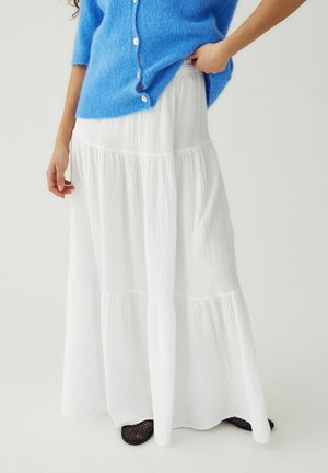 Kobieta ubrana w niebieski miękki kardigan i długą, białą spódnicę warstwową oraz czarne plecione buty, stojąca na jednolitym tle.