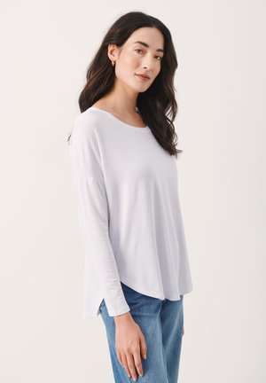 White, long-sleeve top with a round neckline, relaxed fit, and side slits, paired with blue jeans. Smooth texture and lightweight material.