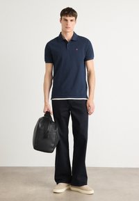 Marine polo skjorte med korte ermer, hvit undertrøye, mørke jeans og en svart lærveske. Beige joggesko fullfører antrekket.