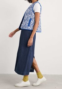 Modrý a bílý květinový vzorovaný vest, kombinovaný s tmavou džínovou sukní a bílými teniskami. Žluté žebrované ponožky viditelné nad botami.