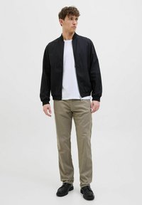 Black bomber jacket with zipper, elastic cuffs, and relaxed fit. Worn over a white t-shirt and beige pants, complemented by black shoes.
