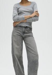 Persona che indossa una maglia grigia a maniche lunghe e jeans grigi larghi, in piedi con le braccia incrociate davanti a uno sfondo uniforme.